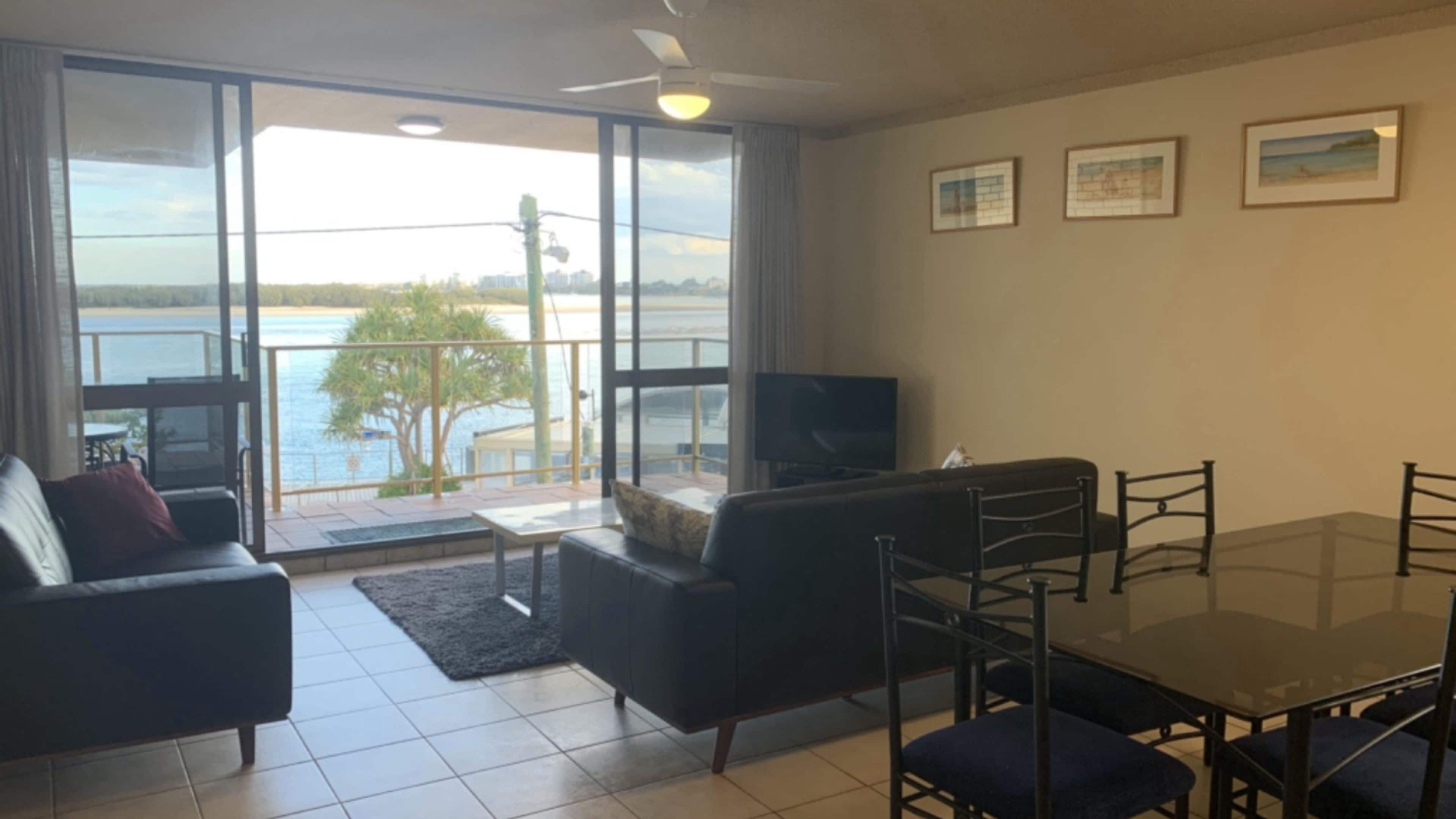 A living room with a sofa, a TV, and a dining table with chairs. There is a balcony with a view of the water.