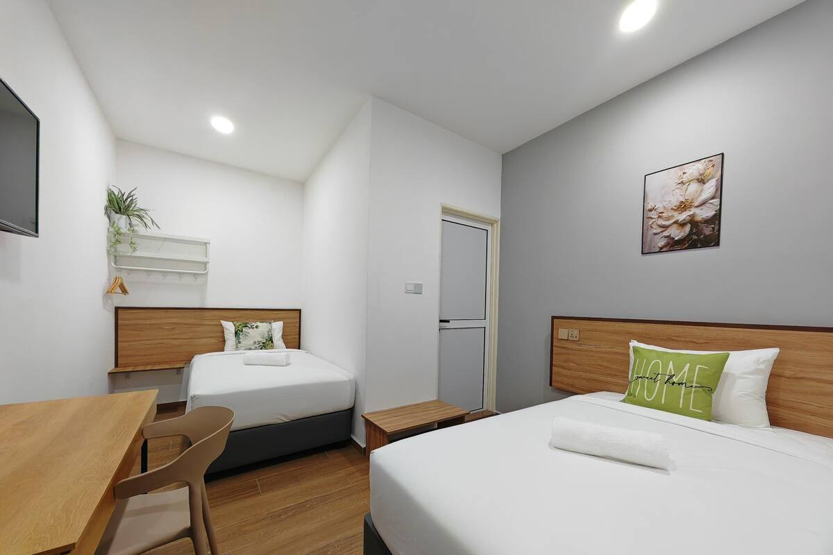 Habitación de hotel con cama, escritorio, silla, una mesita, una planta, una puerta y un cuadro colgado en la pared.