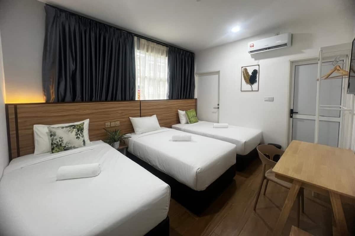 Habitación de hotel con tres camas, cabecera de madera, una mesa, una silla, ventana con cortinas, equipo de aire acondicionado y una puerta.