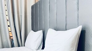 Un lit bien fait, avec du linge de lit blanc et des oreillers, une tête de lit rembourrée de couleur grise et une fenêtre ornée de rideaux beige.