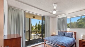 A bedroom with a bed, a nightstand, a wooden dresser, a balcony with a view of trees and a street.
