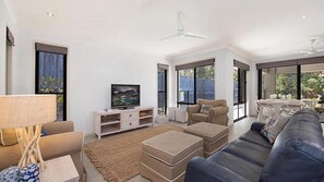 A modern living room with a flat-screen TV, a sofa, ottomans, and a dining area with a table and chairs. - 4BR Pool Hideaway Near Beach (Moffat Beach)