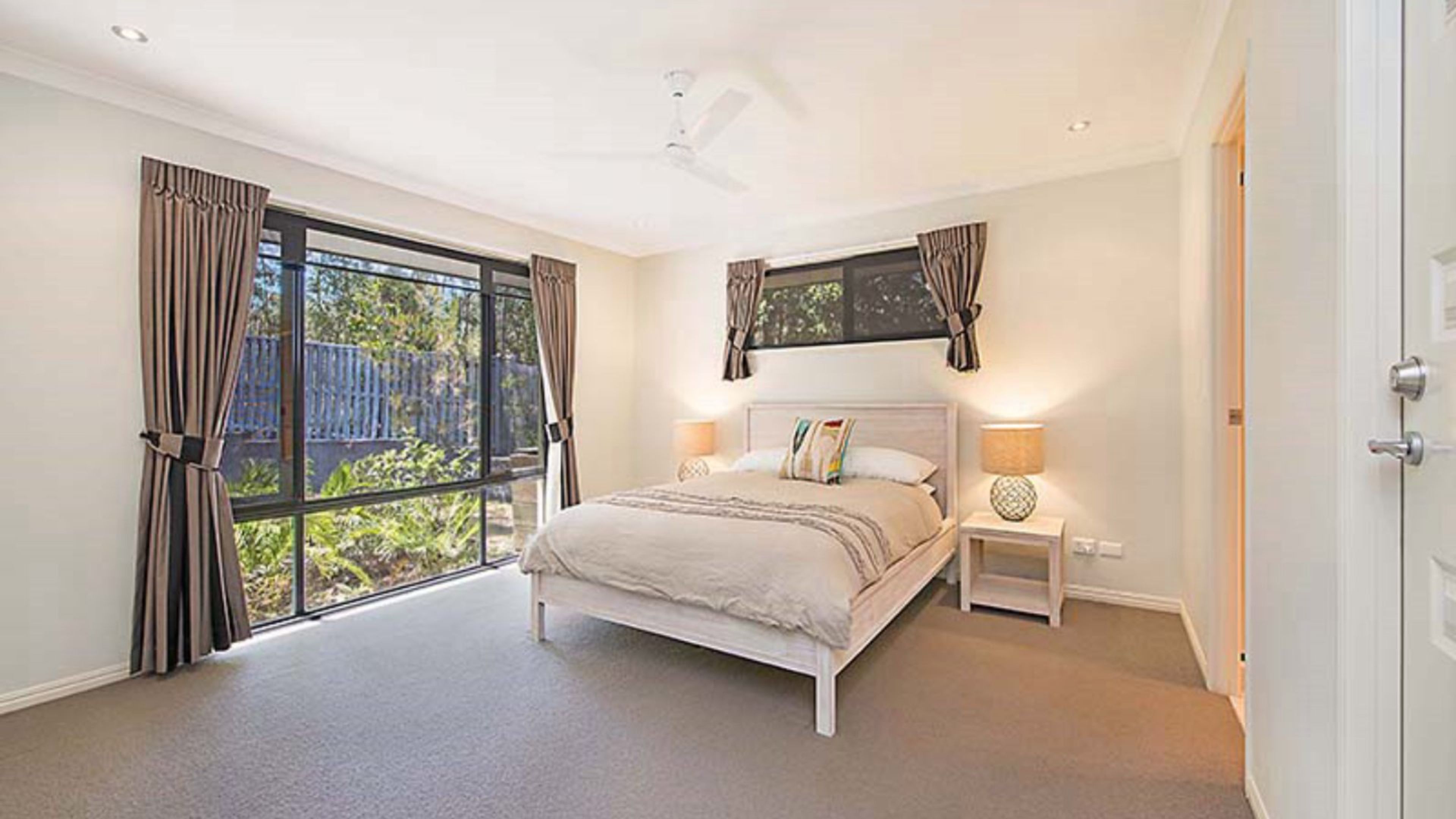 A bedroom with a large window, a bed, a nightstand, and a ceiling fan.