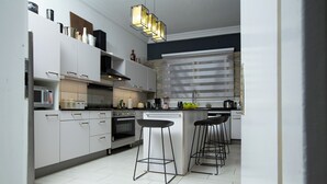 A modern kitchen with white cabinets, a built-in oven, a microwave, and a coffee machine. There are three black bar stools and a large window with blinds.