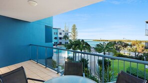 Un balcon avec vue sur une plage et des palmiers, une piscine et un bâtiment aux murs bleus.