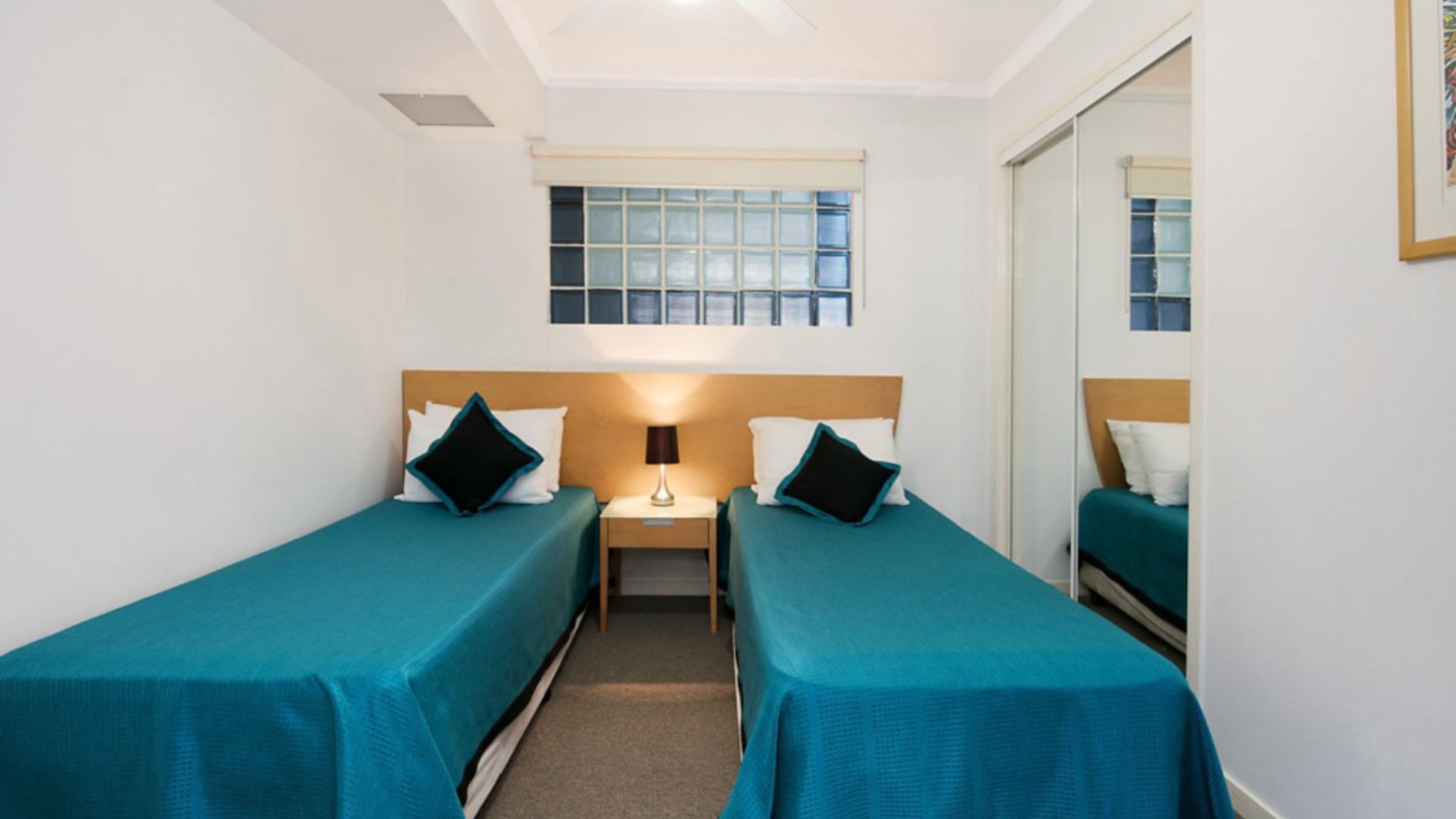Une chambre avec deux lits simples, une table de chevet avec une lampe, une fenêtre avec des stores et un miroir au mur.