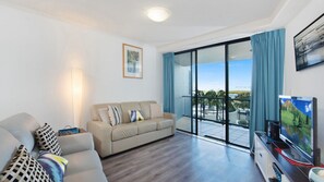 A modern living room with a sofa, a television, and a balcony with a view. - Grand Pacific U19 | Waterfront Views Walk To The Beach (Caloundra)