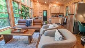Interior acogedor de una cabaña con sala de estar que incluye un sofá y una mesa de centro, cocina equipada con electrodomésticos modernos y ventanas amplias con vistas al bosque.
