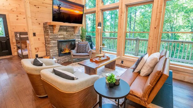 Interior acogedor de una cabaña con chimenea de piedra, asientos cómodos y ventanas amplias con vistas al bosque.