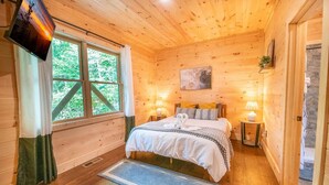 Un dormitorio acogedor tipo cabaña de madera, con cama, dos lámparas de noche, televisión y ventana con cortinas.