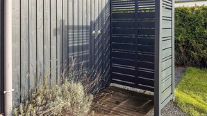 A gated entryway with a wooden fence and a metal gate, leading to a paved area with plants. - 6 Person Holiday Home in Otterup (Otterup)