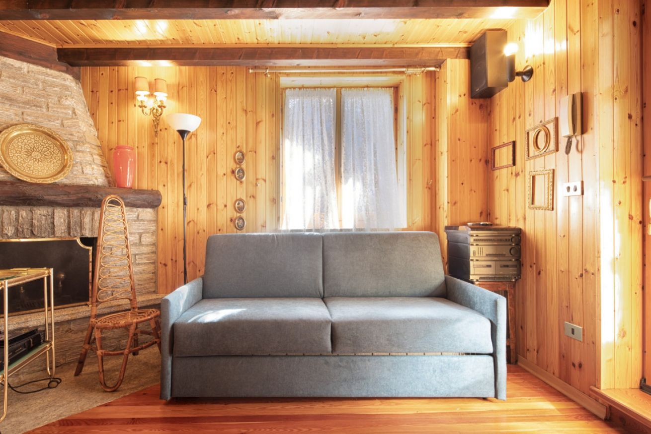 A cozy log cabin interior with a grey sofa, a wicker chair, a stone fireplace, and wooden walls.