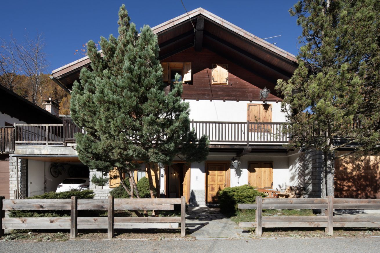 A traditional chalet with a wooden balcony, a stone entrance, and a large pine tree in front.