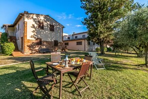 Zona de dinar amb una taula preparada per l'esmorzar, envoltada de cadires, en un ambient exterior assolellat i amb un edifici al fons.