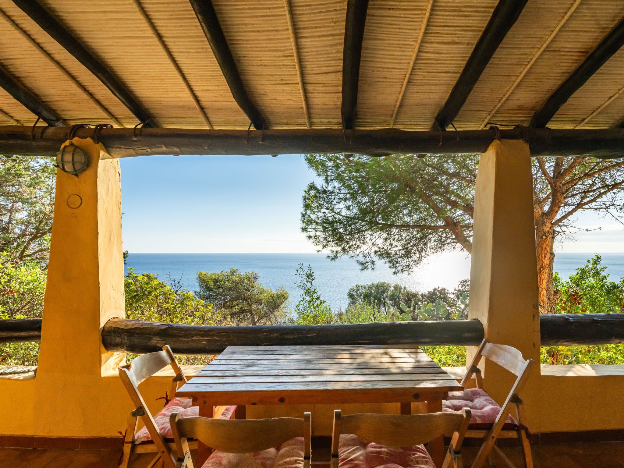 An outdoor wooden table and chairs under a pergola with a view of the sea.