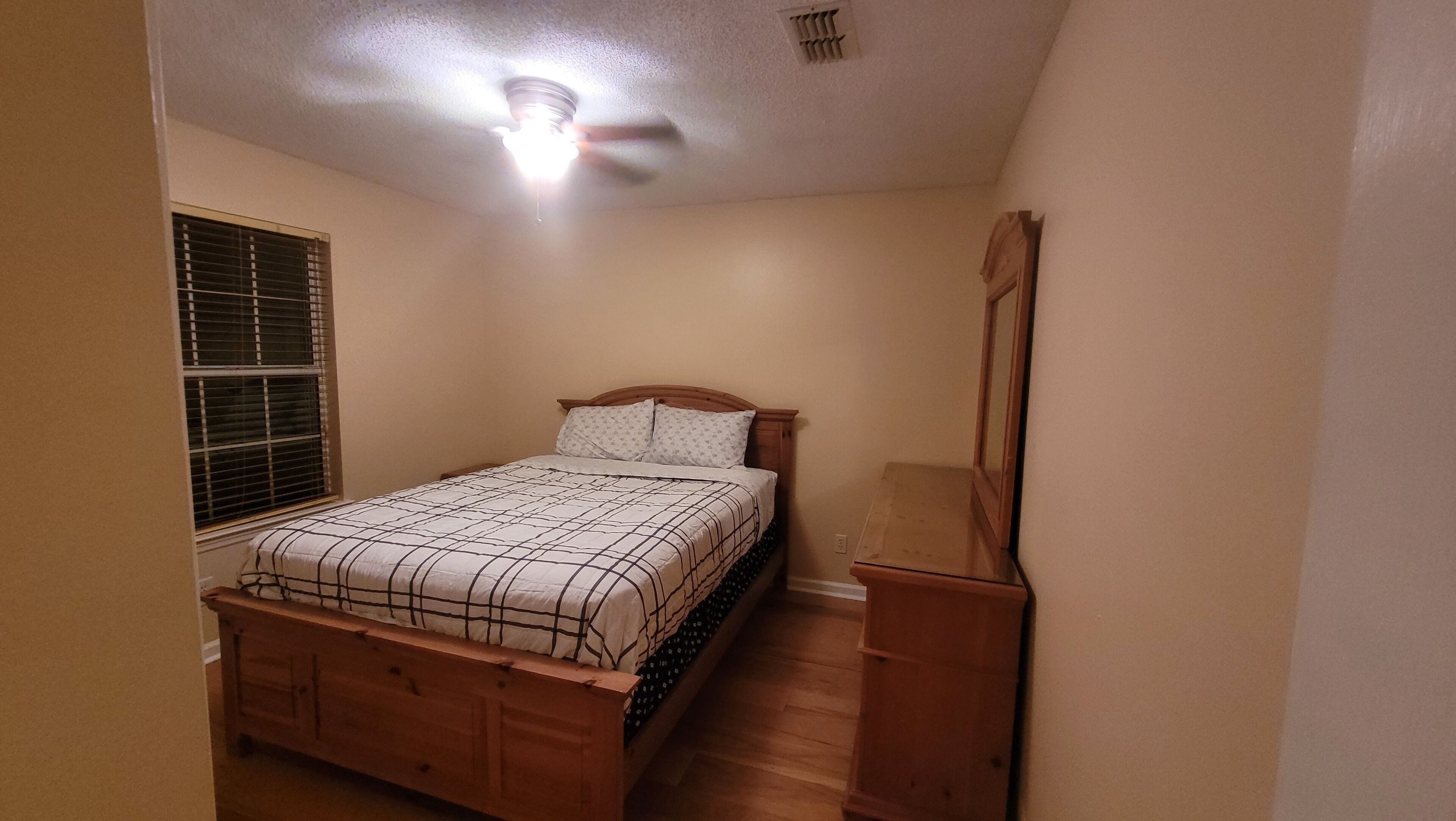 Une chambre à coucher avec un lit, une commode, une fenêtre avec des stores et un ventilateur de plafond.