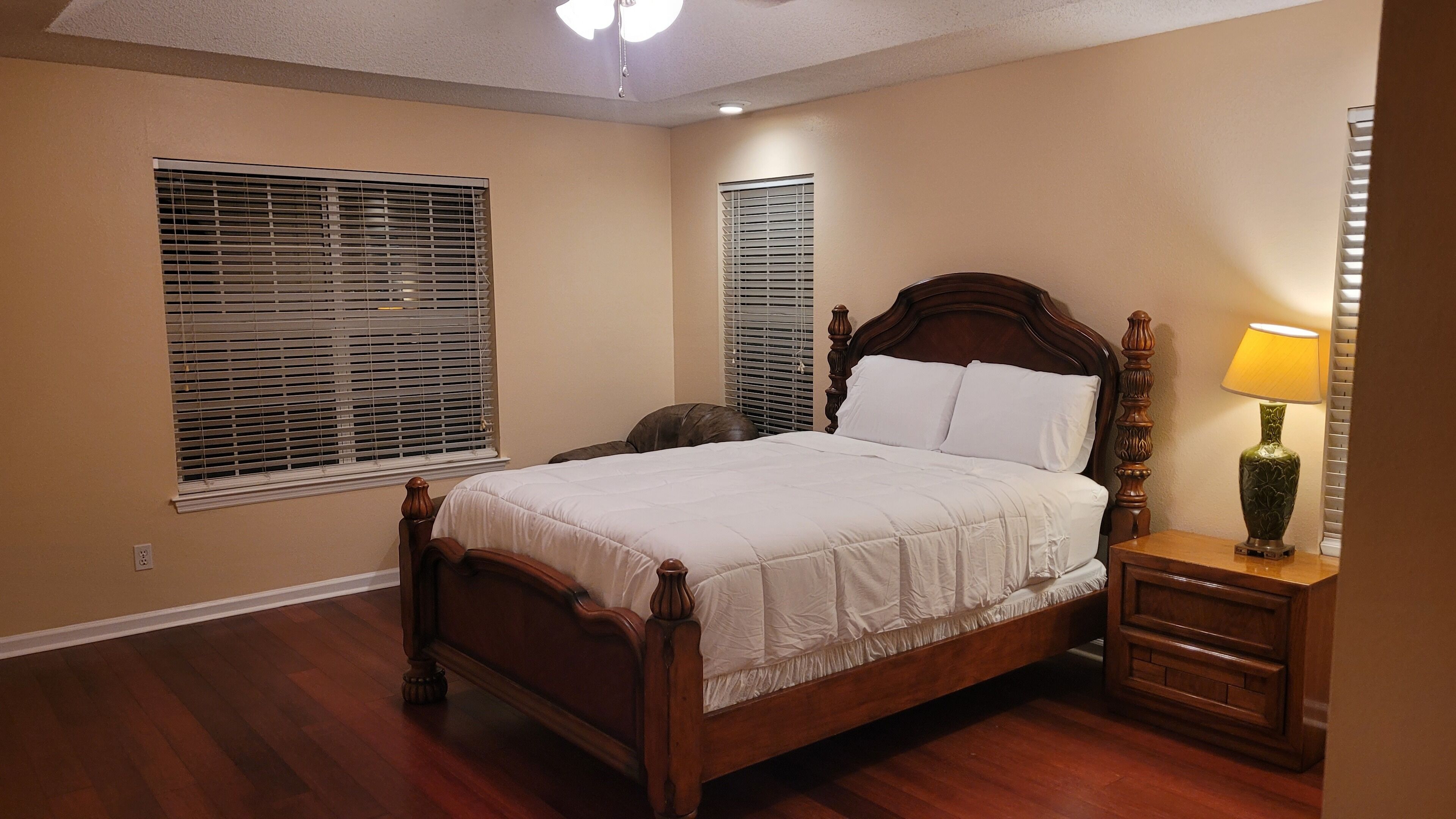 A bedroom with a wooden bed, two windows with blinds, a nightstand with a lamp, and a wooden floor.