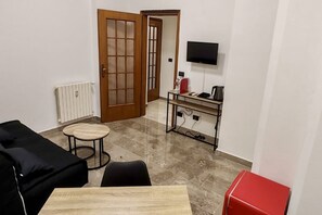 Une pièce avec un canapé noir, une petite table ronde, un meuble rouge, un téléviseur fixé au mur et une porte donnant sur une autre pièce.