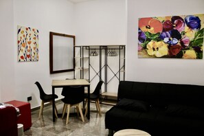 Une pièce avec un canapé noir, une table en bois avec des chaises, une valise rouge et deux œuvres d’art accrochées aux murs.