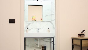 A bathroom with a marble sink, a mirror, a shelf with toiletries, and a small table with a remote control. - Arsenale Suites, Love Nest. A cozy retreat for two, where romance meets comfort (Torino)