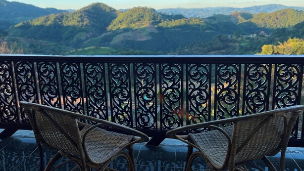 Un balcon doté de chaises en osier et d’une rambarde en métal décorative, offrant une vue sur un paysage montagneux.