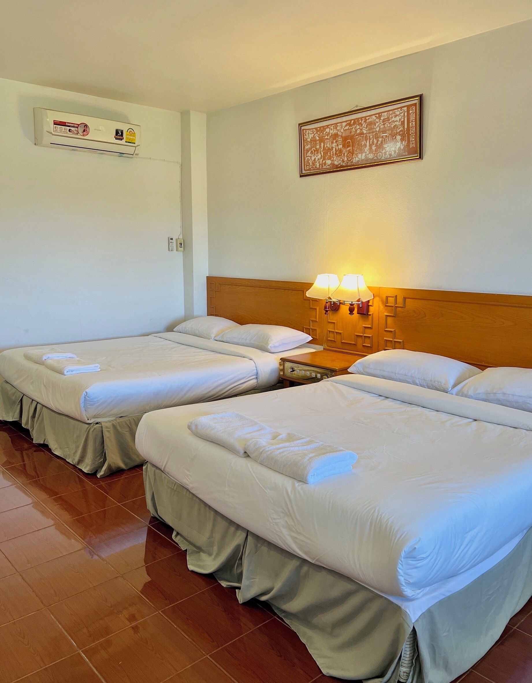 A hotel room with two single beds, a wooden headboard, a wall-mounted air conditioner, a framed picture, and a bedside lamp.