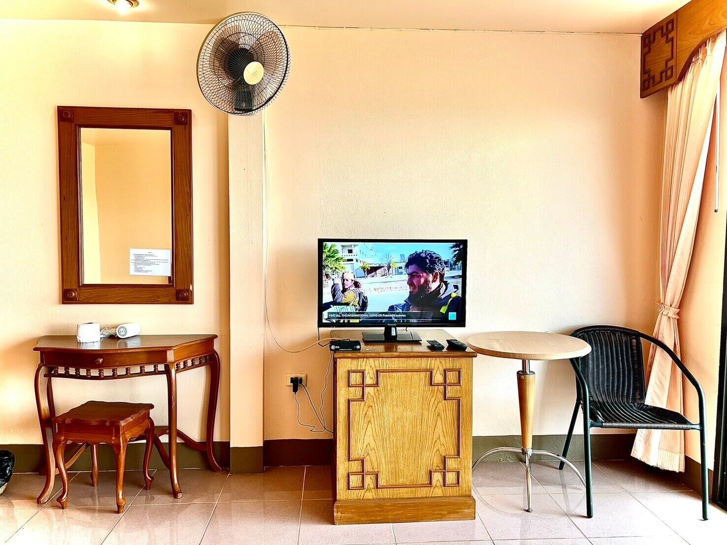 A hotel room with a wooden desk, a small chair, a television on a wooden cabinet, a standing fan, and a window with curtains.