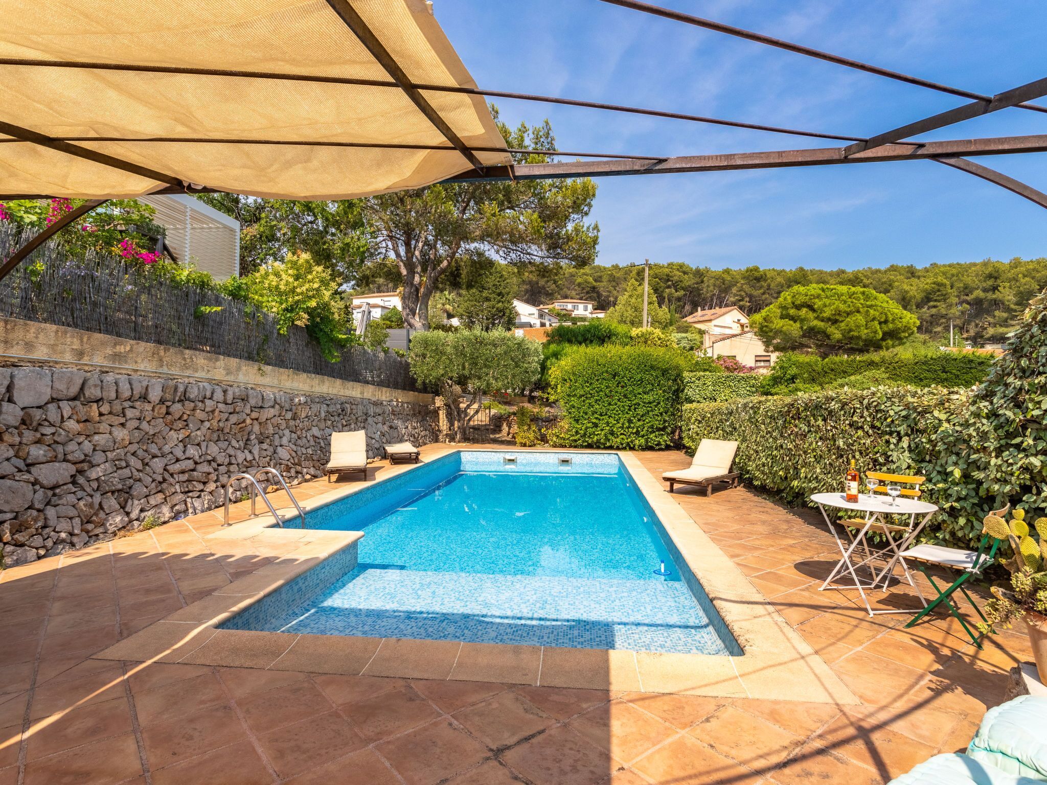 A rectangular swimming pool with a tiled edge, surrounded by a stone wall and a tiled patio. There are lounge chairs and a table with a bottle and glasses.