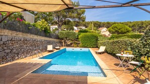 A rectangular swimming pool with a tiled edge, surrounded by a stone wall and a tiled patio. There are lounge chairs and a table with a bottle and glasses.