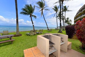 A beachside patio with a grill, lounge chairs, and palm trees. - Direct Oceanfront Views Remodeled 2BD Condo-Paki Maui 301 (Lahaina)