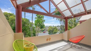 A balcony with a view of palm trees and a red car on the street. There are two chairs, one green and one red.
