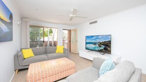 A modern living room with a grey sofa, a patterned ottoman, a television on a white stand, and a ceiling fan.