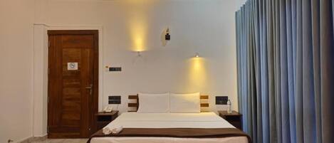 A hotel room with a large bed, a wooden door, a wall-mounted light, and a curtain.