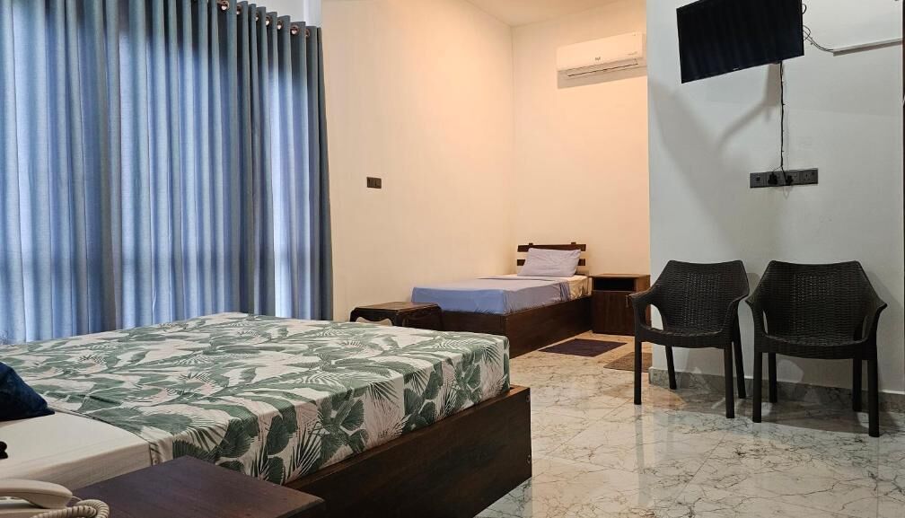 A hotel room with a bed, a desk with a telephone, two wicker chairs, a TV mounted on the wall, and a window with curtains.
