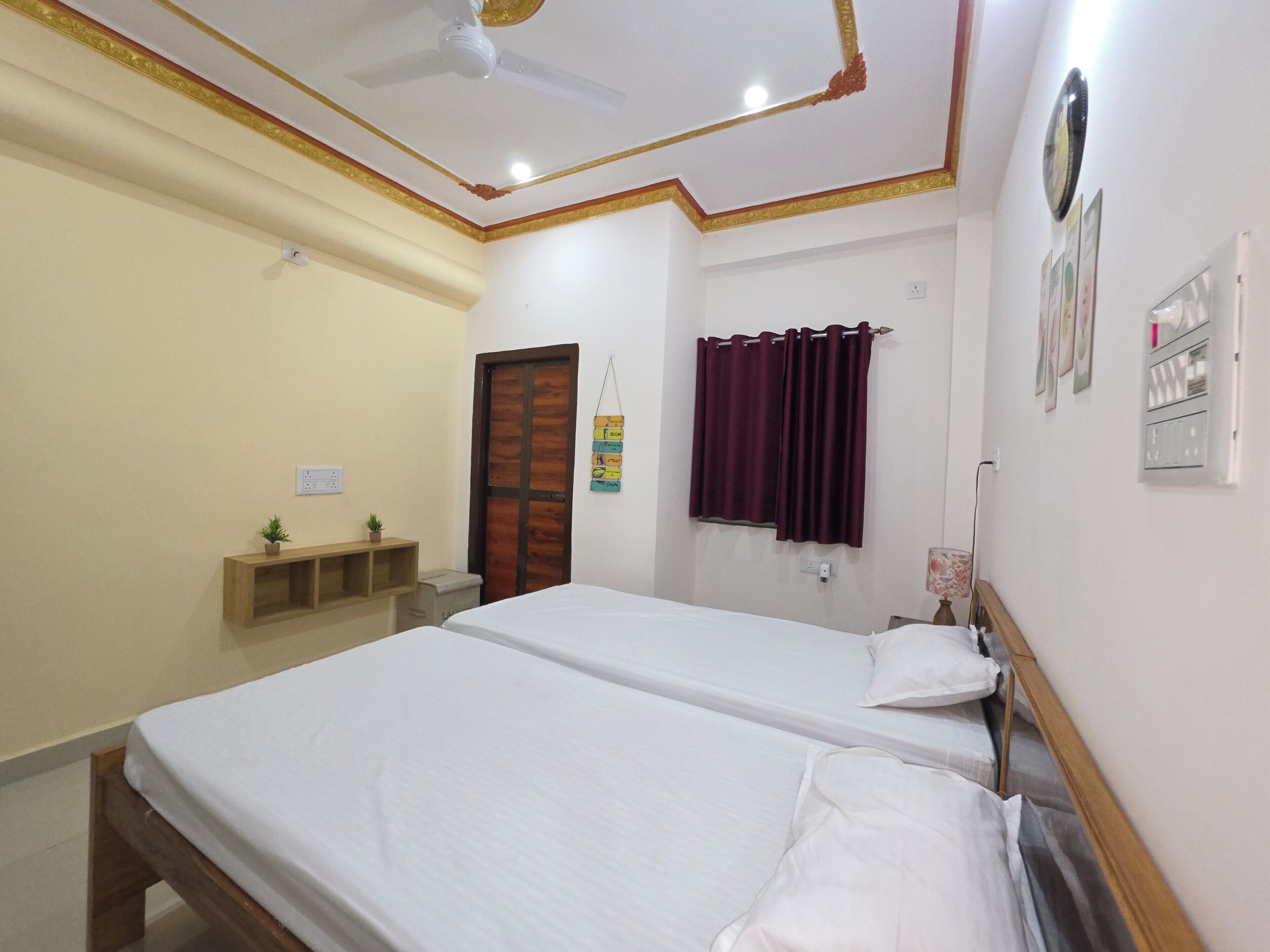 Una habitación de hotel con dos camas individuales, un ventilador de techo, una puerta de madera, una ventana con cortinas, un estante pequeño con plantas y un reloj fijado a la pared.