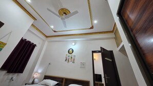A hotel room with a ceiling fan, a bed with two pillows, a nightstand with a lamp, a wall clock, and a door with a mirror. - Narayans Residency (Gaya)