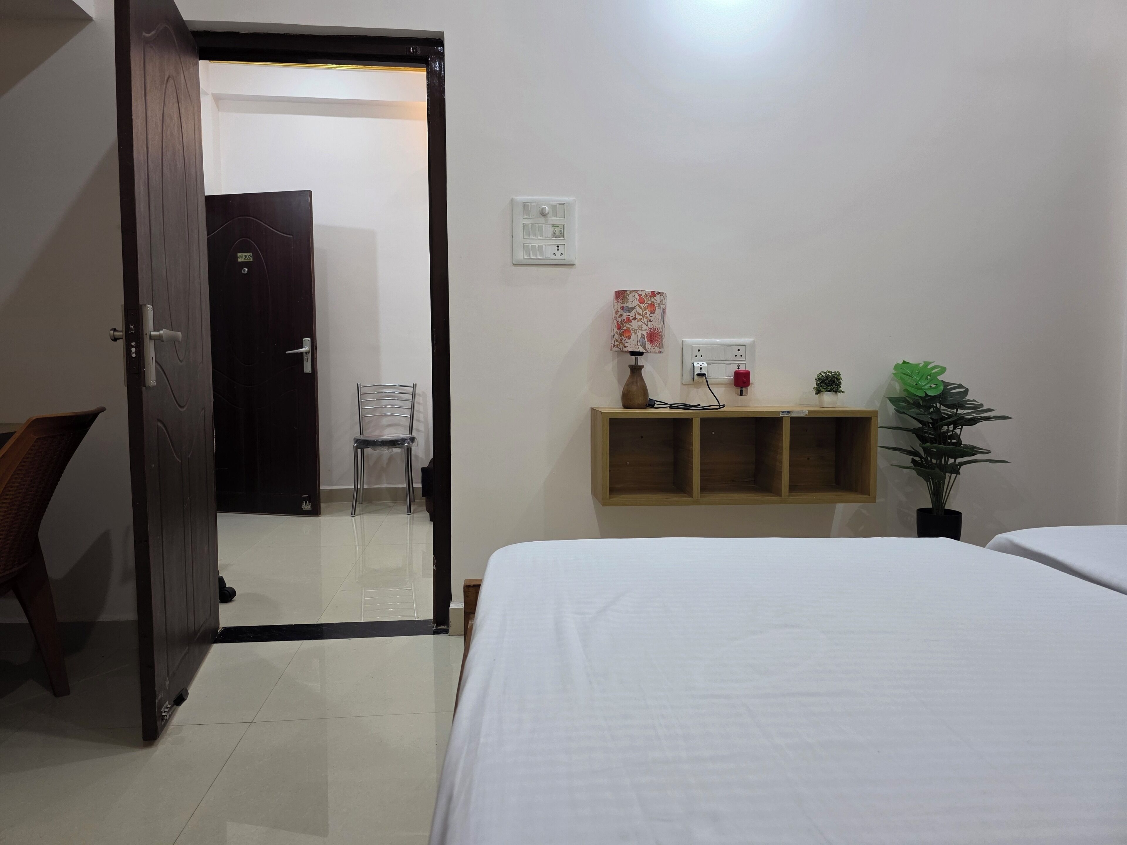 A hotel room with a bed, a wooden shelf with a lamp, a chair, and a potted plant.