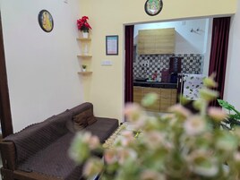 Una sala de estar con un sofá marrón, una pequeña estatua blanca y una flor roja sobre una estantería. Se ve una cocina a través de un umbral abierto, con armarios de madera y un salpicadero azulejado.