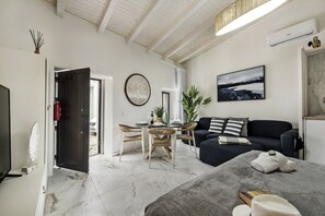 A modern living room with a dark sofa, a dining table with chairs, a TV, and a large window.