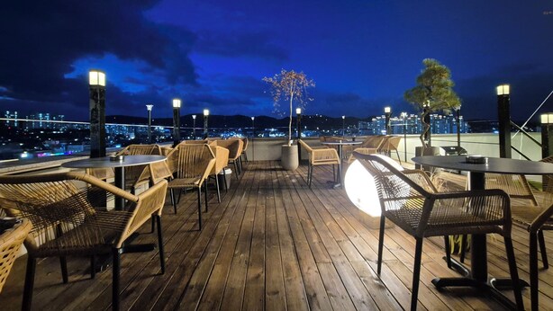 An outdoor seating area with wicker chairs and tables, wooden flooring, and city lights in the background. - marihotel (Seosan)