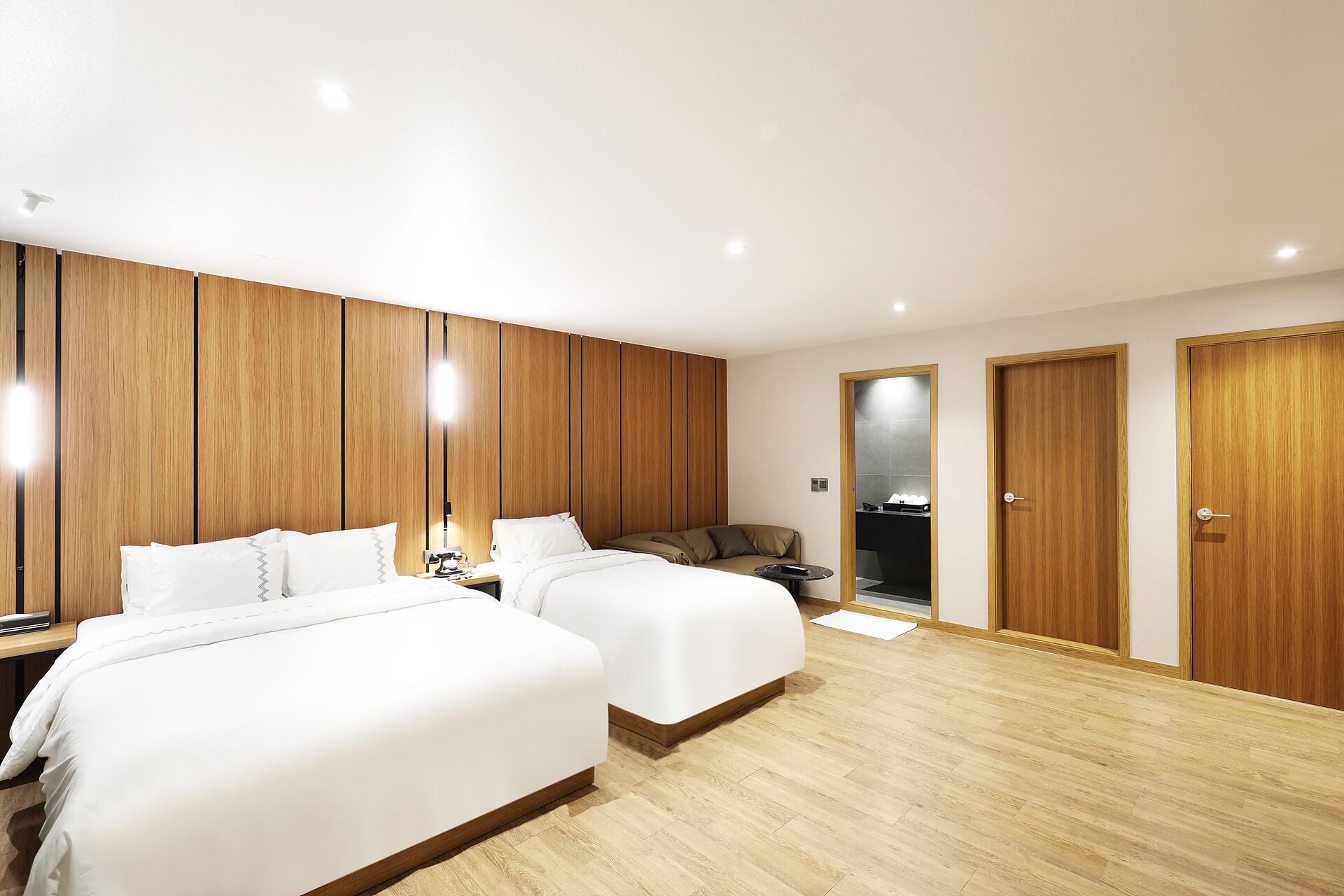 A hotel room with two beds, wooden paneling, a small seating area, and a bathroom visible through an open door.