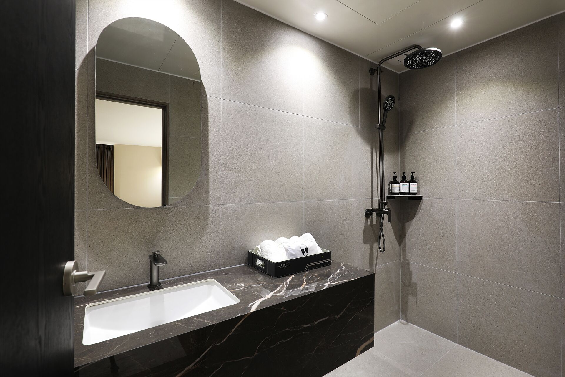 A modern bathroom with a marble sink, a large mirror, a shower, and toiletries on a shelf.