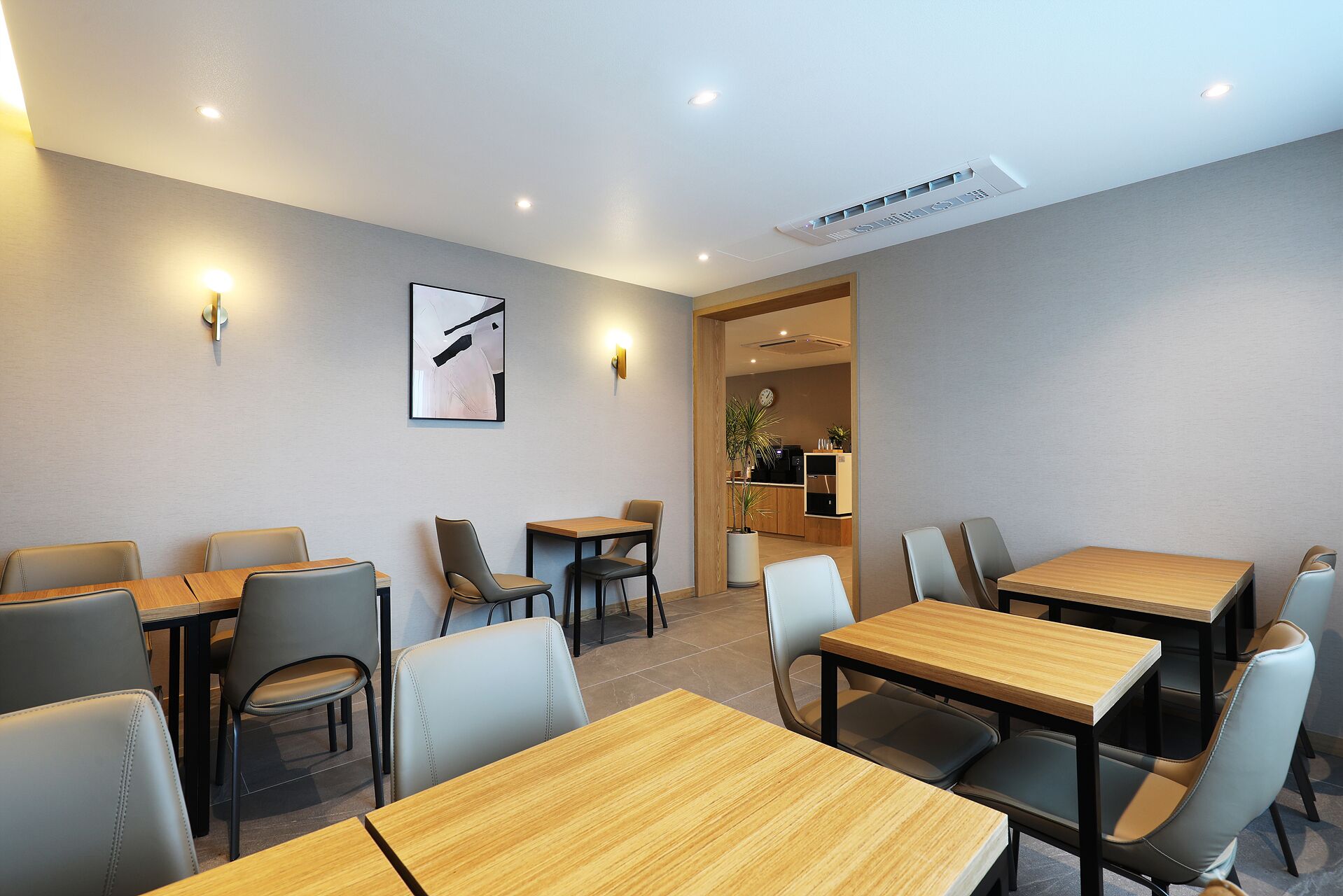 A modern dining area with wooden tables and chairs, a clock on the wall, and a kitchen visible through an open doorway.