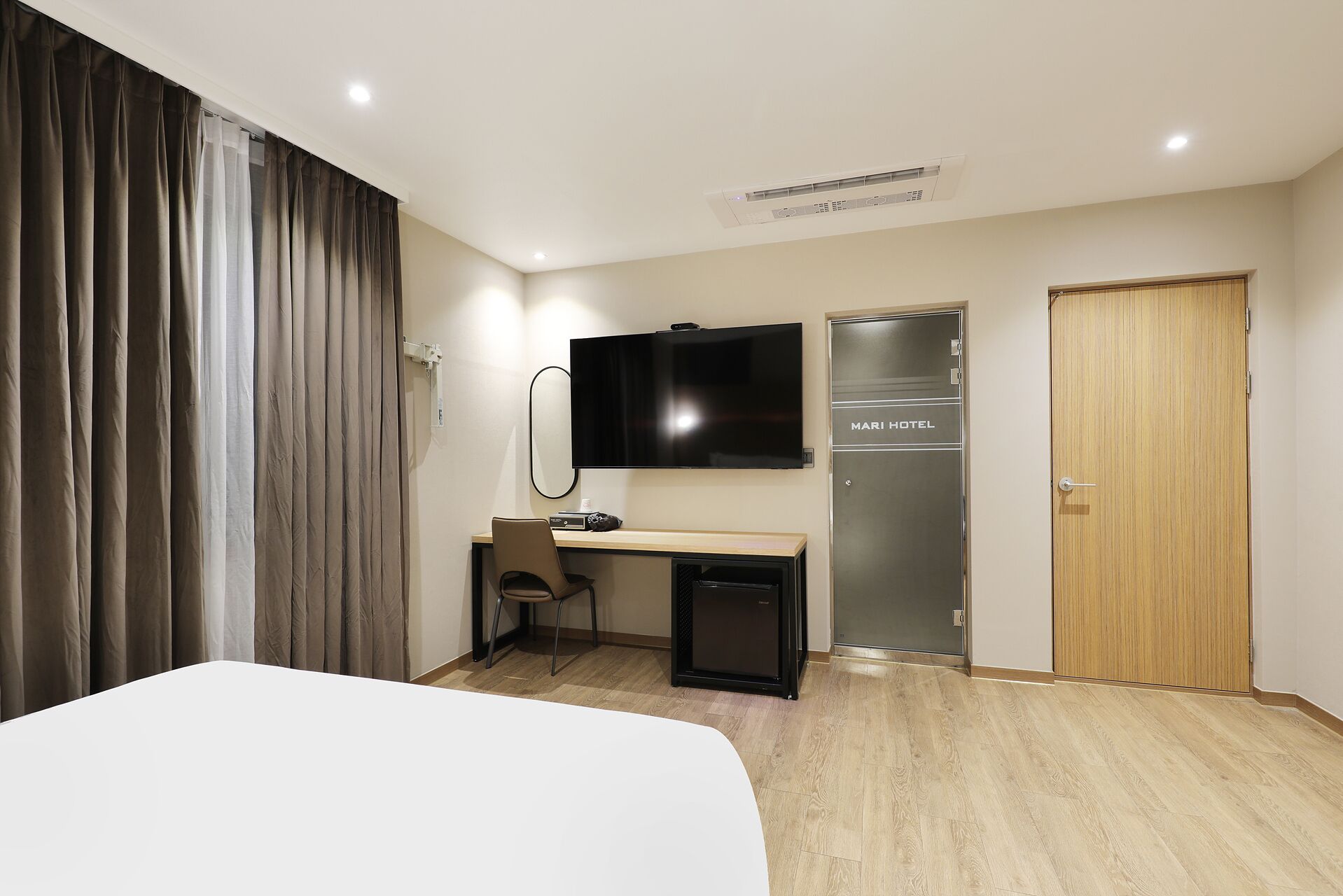 Ett hotellrum med en säng, ett skrivbord med en stol, en TV, en minibar och en dörr med texten ”MARI HOTEL”.