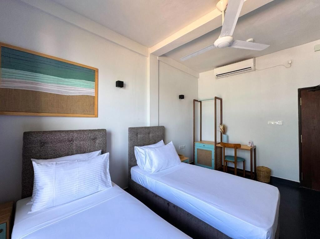 A hotel room with two single beds, a ceiling fan, an air conditioning unit, a small desk with a chair, and a mirror.
