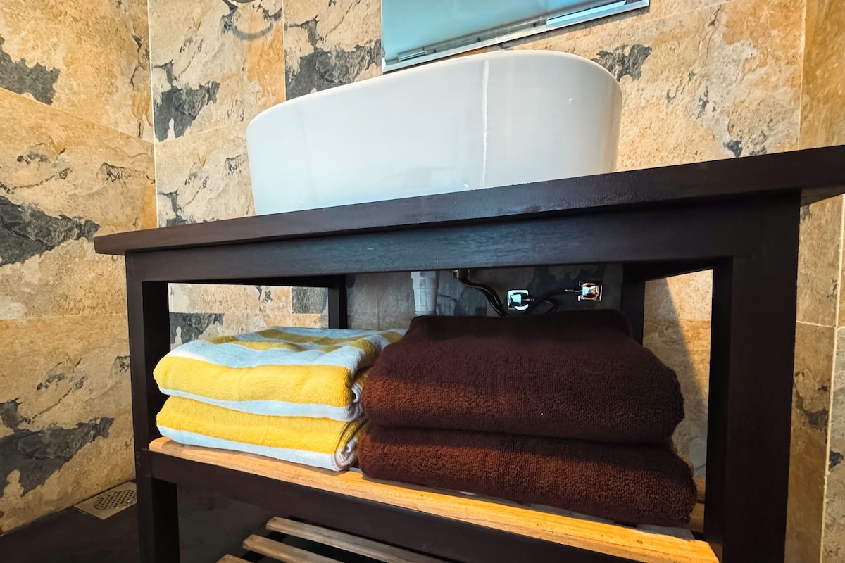 Un mobile bagno con un lavandino bianco, un porta asciugamani con asciugamani gialli e marroni, e uno scaffale in legno.