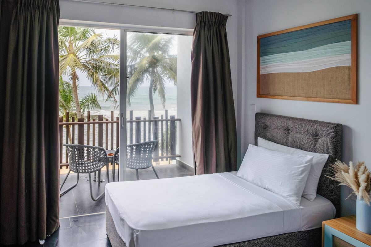 Een hotelkamer met een bed, uitzicht op de oceaan, een balkon met stoelen en een schilderij aan de muur.