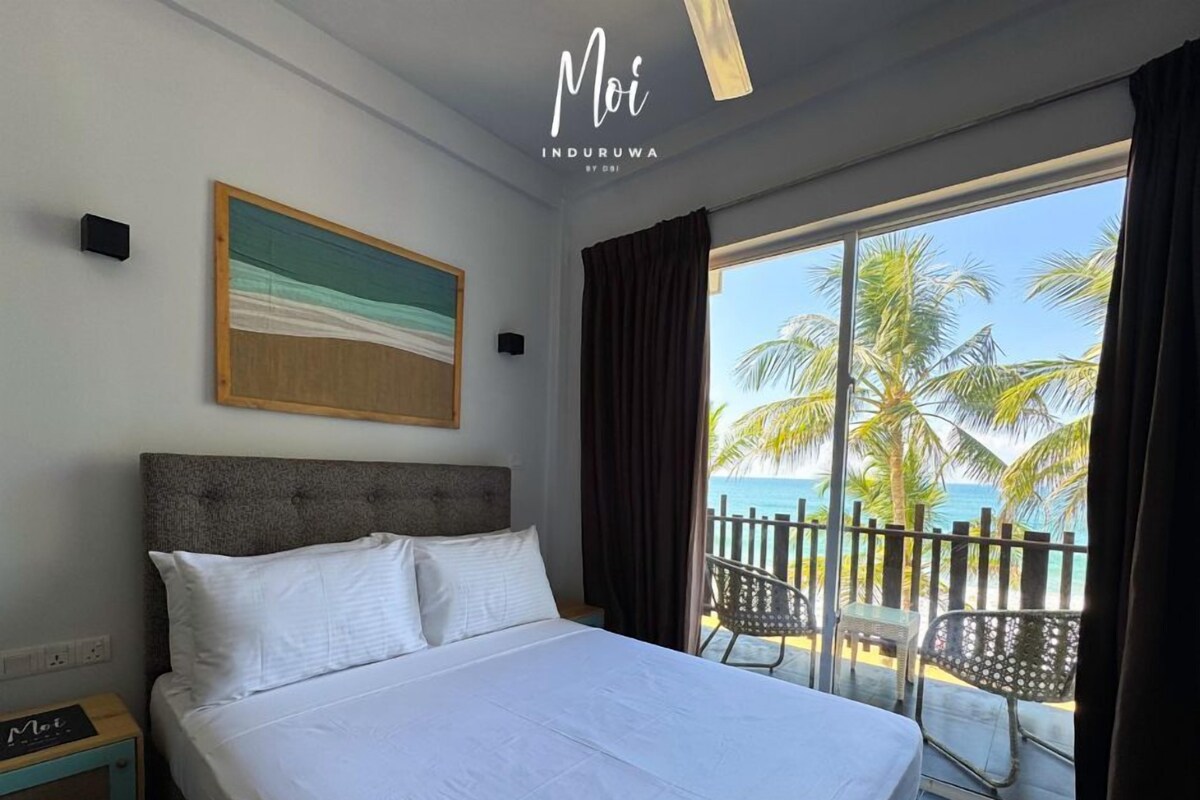 A hotel room with a bed, a painting, a view of the beach, and a balcony with chairs.