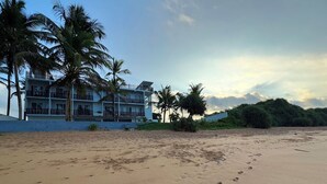 A beach with footprints leading to a building with balconies and palm trees. - Moi Induruwa by DBI (Induruwa)