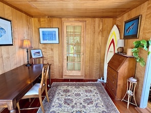 A wooden-paneled room with a wooden desk, chairs, a rug, a dresser, a surfboard, and framed pictures on the walls. - Lillie’s Oceanview RUSTIC Boho Surf Cottage
Steps to the beach & fun! (Summerland)